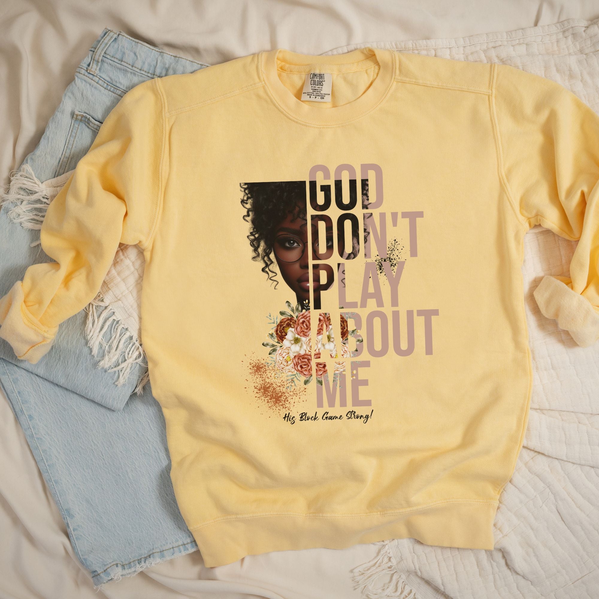 Flatlay mockup of women's butter color sweatshirt with design that reads "God Don't Play About Me - His Block Game Strong" and dark skin lady graphic