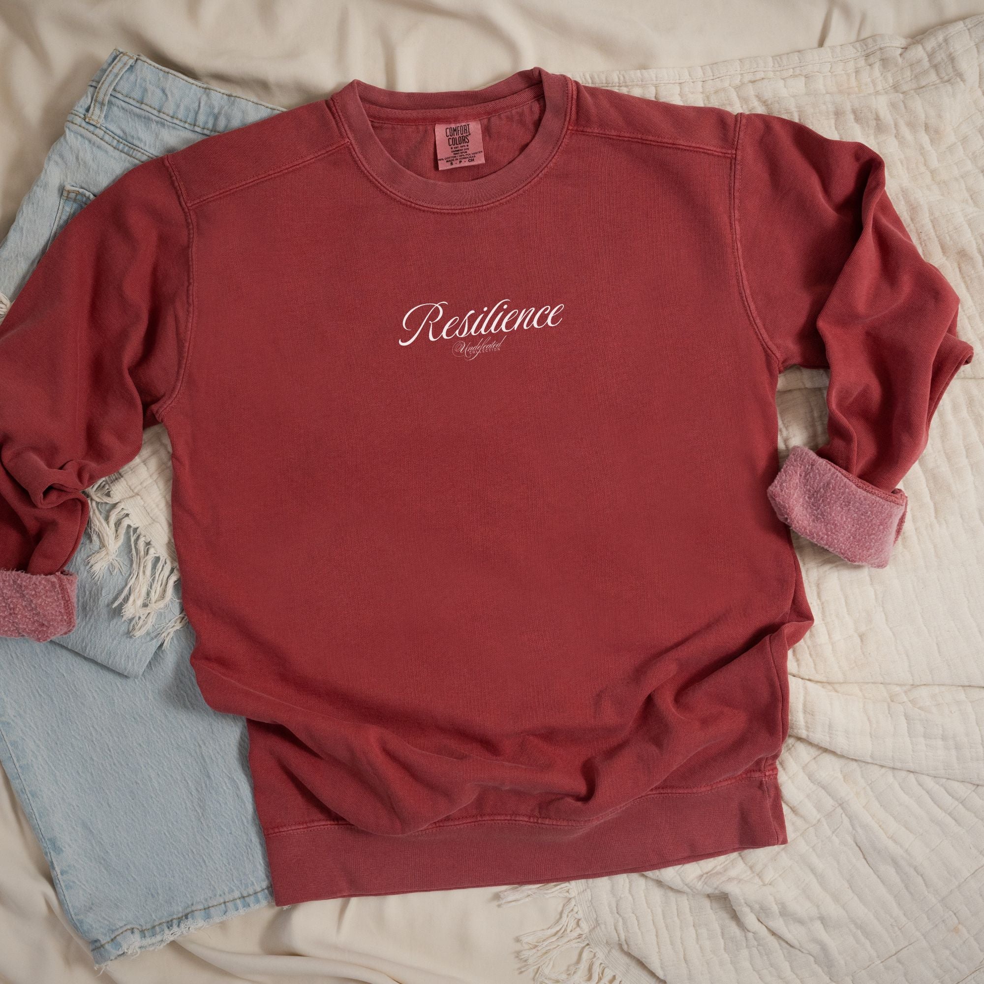 A Flat lay mockup of front design of crimson color sweatshirt with text design that partly reads "Resilience - Undefeated"